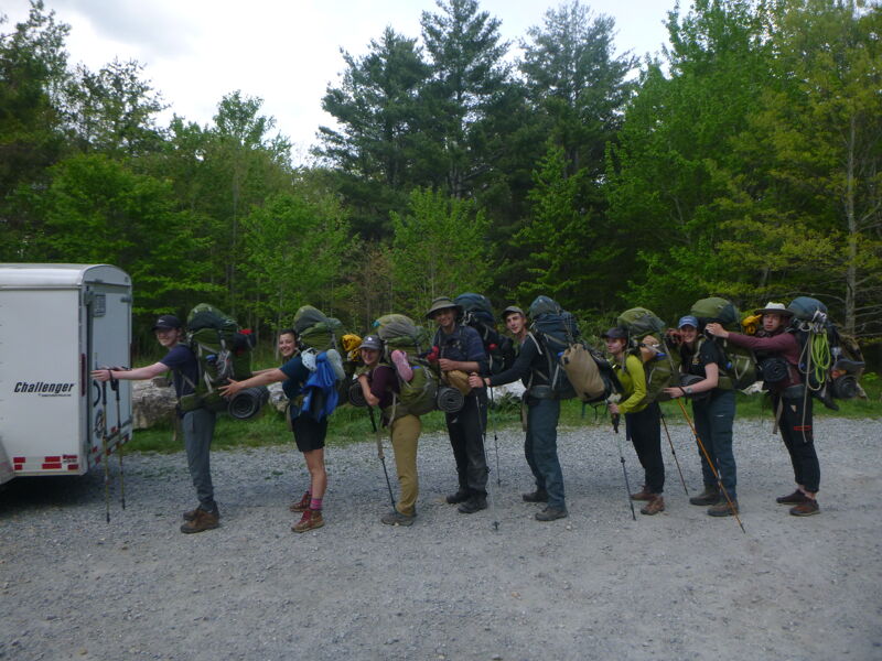 A group of about ten people stand in a line on a gravel road, each wearing large backpacks and holding hiking poles. They appear to be preparing for a hike, possibly unloading gear from a small trailer parked nearby. The background features a dense forest, suggesting a wilderness or outdoor recreational area.